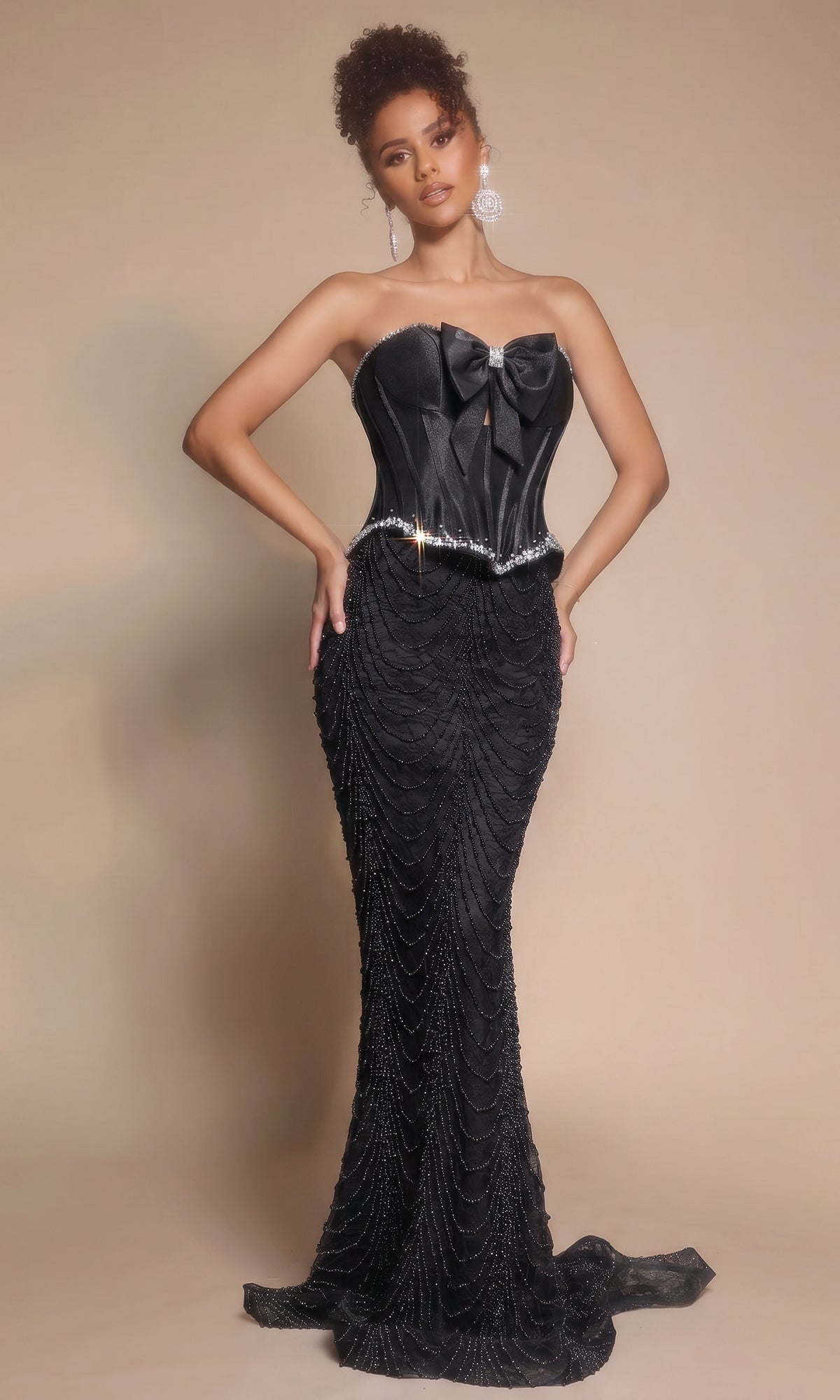 A woman poses against a neutral background in the Portia and Scarlett PS26054 long black formal dress, featuring a strapless corset bodice, bow accents, and a beaded fitted skirt.